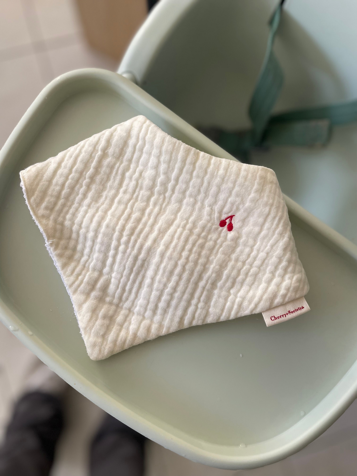 Cherry + Society vanilla muslin bandana bib with delicate red cherry embroidery on a high chair tray